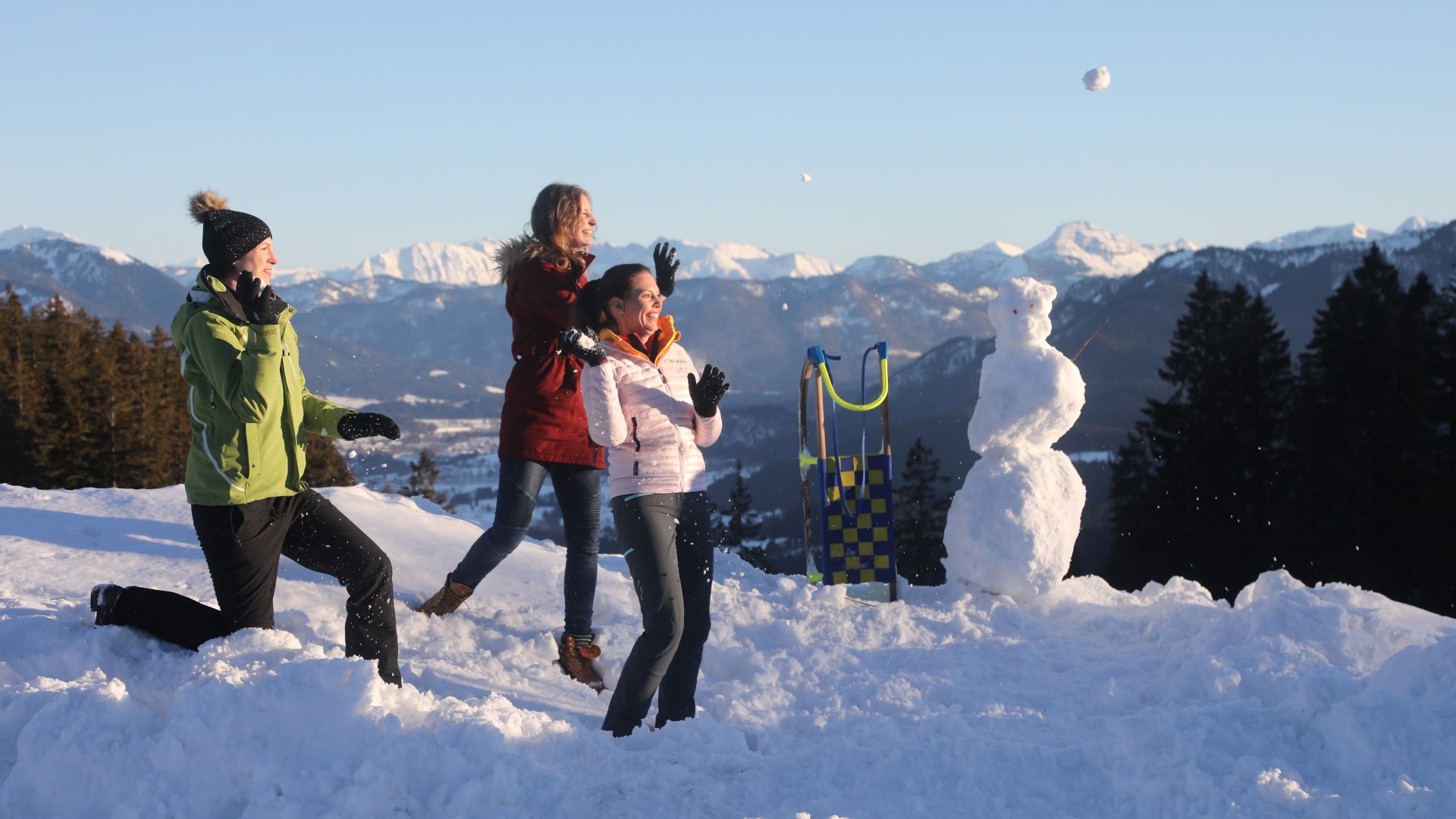 Das Bild zeigt drei Frauen in Winterkleidung, die Schnebälle in Richtung eines Rodels und Schneemannes werfen. Im Hintergrund sieht man die winterliche Bergkulisse, © Tourist Information Bad Tölz Foto: Hias Krinner Das Bild zeigt drei Frauen in Winterkleidung, die Schnebälle in Richtung eines Rodels und Schneemannes werfen. Im Hintergrund sieht man die winterliche Bergkulisse, © Tourist Information Bad Tölz Foto: Hias Krinner