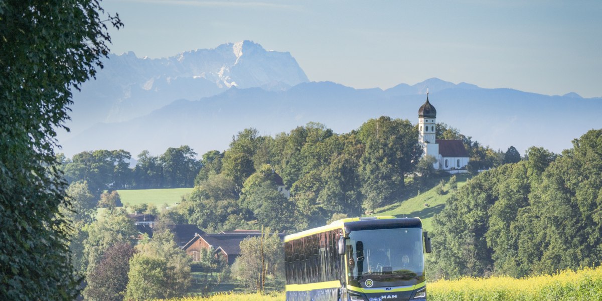 Der Bus fährt durch die hügelige Landschaft, im Hintergrund die Kirche von Holzhausen/Münsing und die Bergkette mit der markanten Zugspitze, © Landratsamt Bad Tölz-Wolfratshausen|Dietmar Denger Der Bus fährt durch die hügelige Landschaft, im Hintergrund die Kirche von Holzhausen/Münsing und die Bergkette mit der markanten Zugspitze, © Landratsamt Bad Tölz-Wolfratshausen|Dietmar Denger