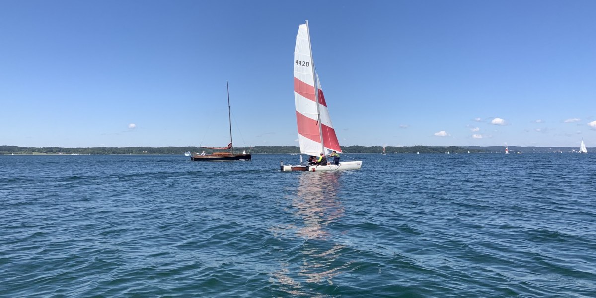 auf dem blauen Wasser des Starnberger Sees segelt ein Katamaran mit Rot-weißem Segel, © Wassersportcenter Starnberger See, Foto: Wallusch auf dem blauen Wasser des Starnberger Sees segelt ein Katamaran mit Rot-weißem Segel, © Wassersportcenter Starnberger See, Foto: Wallusch