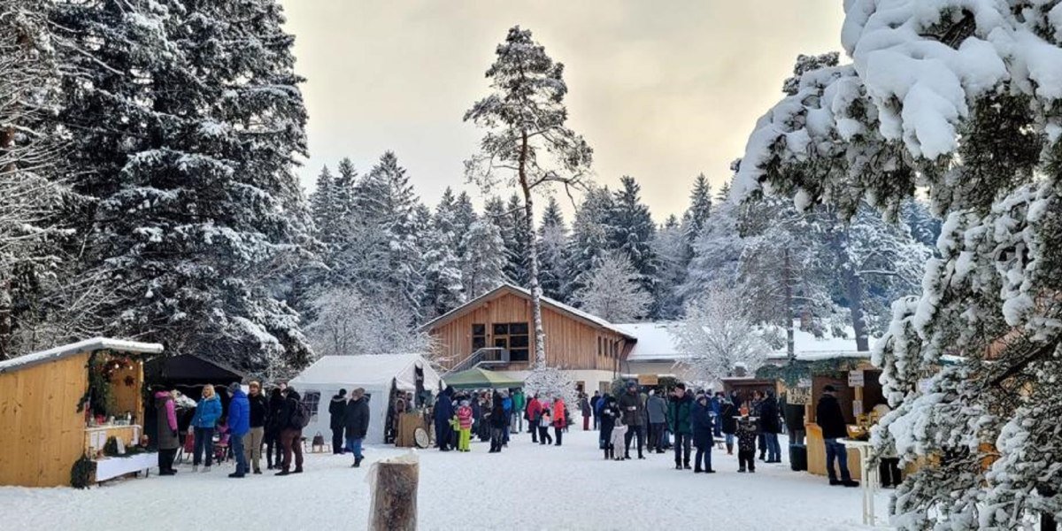 Das Bild zeigt einen winterlichen Weihnachtsmarkt im Wald mit mehreren Ständen und vielen Besuchern, © Jugendsiedlung Hochland, Königsdorf Das Bild zeigt einen winterlichen Weihnachtsmarkt im Wald mit mehreren Ständen und vielen Besuchern, © Jugendsiedlung Hochland, Königsdorf