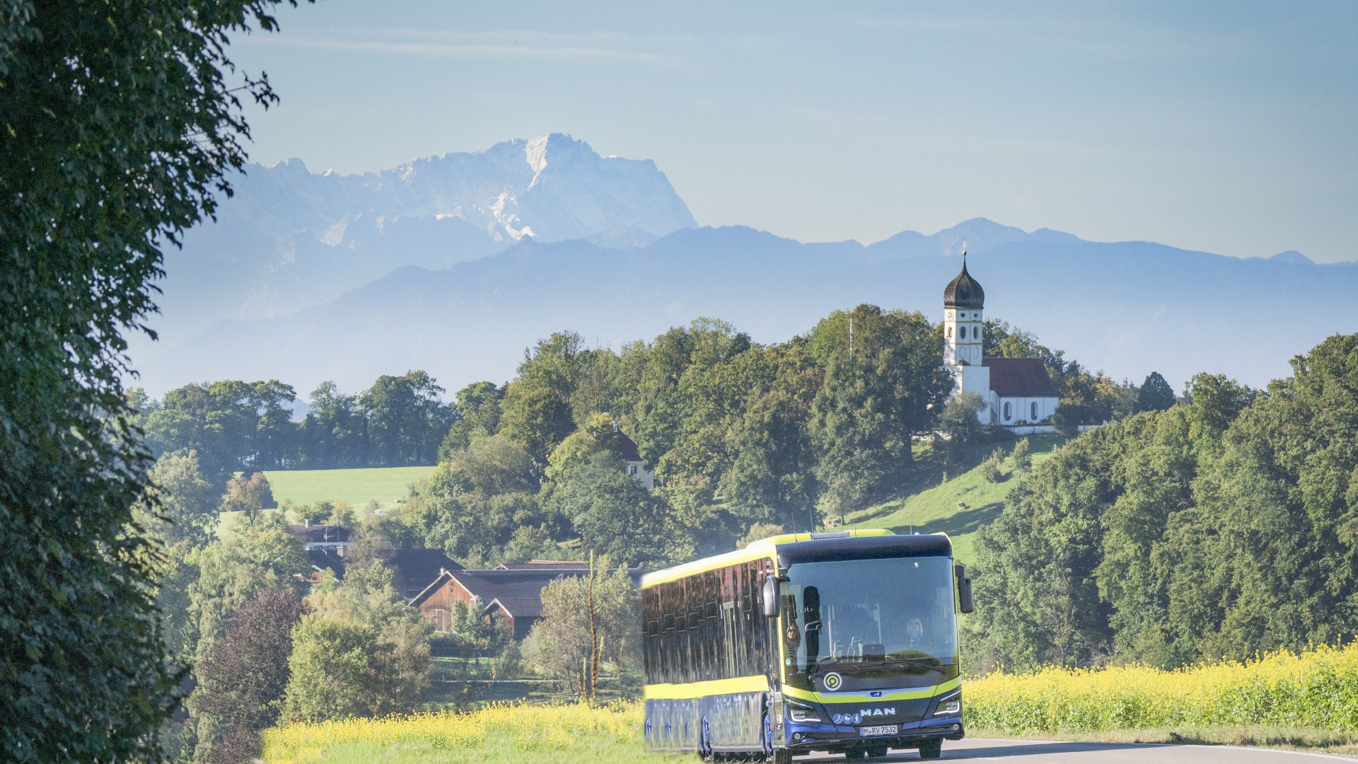 Der Bus fährt durch die hügelige Landschaft, im Hintergrund die Kirche von Holzhausen/Münsing und die Bergkette mit der markanten Zugspitze, © Landratsamt Bad Tölz-Wolfratshausen|Dietmar Denger Der Bus fährt durch die hügelige Landschaft, im Hintergrund die Kirche von Holzhausen/Münsing und die Bergkette mit der markanten Zugspitze, © Landratsamt Bad Tölz-Wolfratshausen|Dietmar Denger