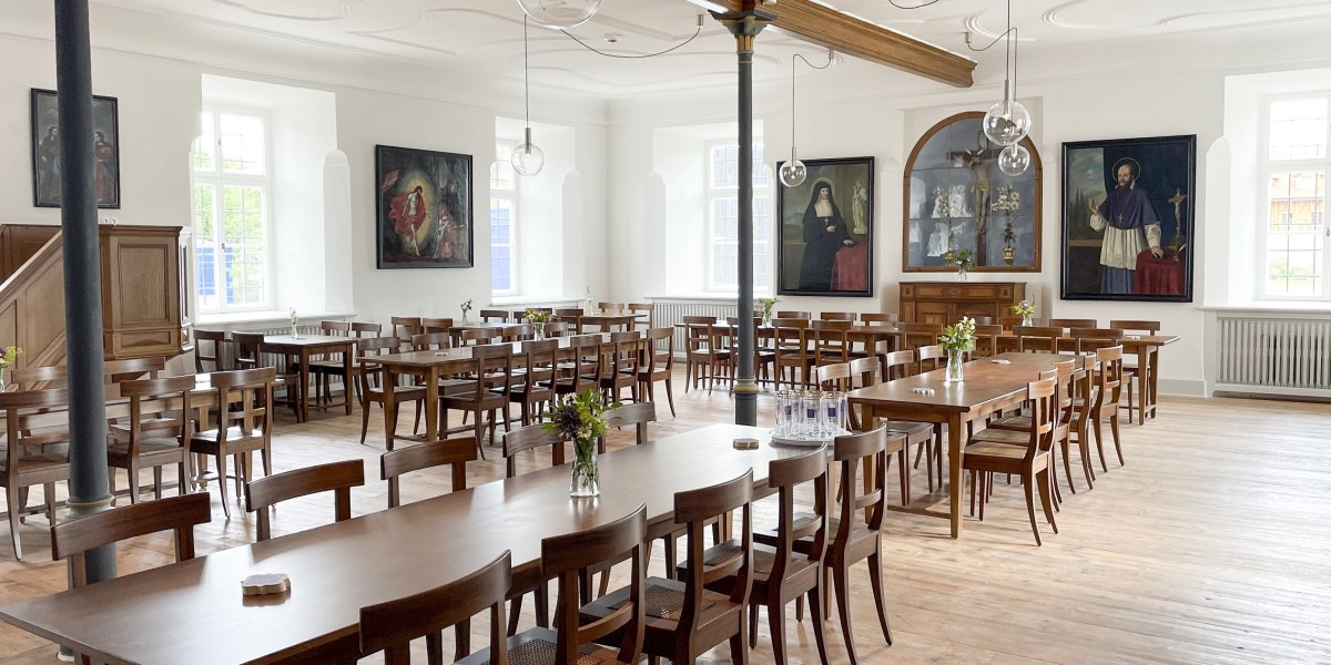 Hell und freundlich - der Restaurantbereich im Kloster Beuerberg.
Das Bild zeigt einen hellen Speisesaal mit Holzboden und langen Holztischen, umgeben von vielen Stühlen. Auf den Tischen stehen Vasen mit frischen Blumen. Große Fenster lassen Tageslicht herein. An den weißen Wänden hängen vier große Gemälde mit religiösen Motiven., © Kloster Beuerberg|Foto: Christian Schmid Hell und freundlich - der Restaurantbereich im Kloster Beuerberg.
Das Bild zeigt einen hellen Speisesaal mit Holzboden und langen Holztischen, umgeben von vielen Stühlen. Auf den Tischen stehen Vasen mit frischen Blumen. Große Fenster lassen Tageslicht herein. An den weißen Wänden hängen vier große Gemälde mit religiösen Motiven., © Kloster Beuerberg|Foto: Christian Schmid