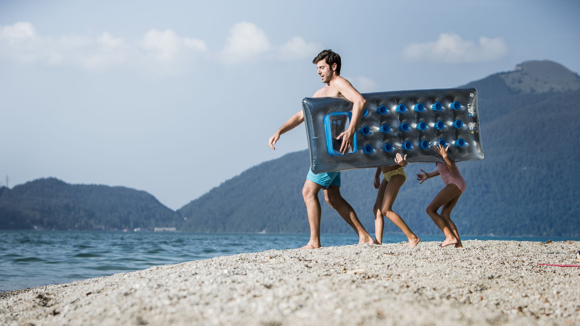 Ausgerüstet mit einer Luftmatratze spaziert ein Mann mit zwei kleinen Kindern am Strand des Walchensees in Richtung Wasser, © Tölzer Land Tourismus|H. Heckmair Ausgerüstet mit einer Luftmatratze spaziert ein Mann mit zwei kleinen Kindern am Strand des Walchensees in Richtung Wasser, © Tölzer Land Tourismus|H. Heckmair