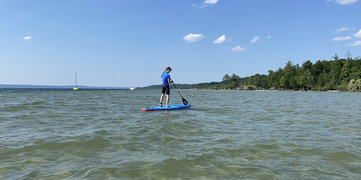 Ein blau gekleideter Wassersportler steht auf einem blauen SUP-Board und paddelt durch den Starnberger See bei Münsing. Im Hintergrund sind einige Segelboote zu sehen., © Wassersportcenter Starnberger See, Foto: Wallusch Ein blau gekleideter Wassersportler steht auf einem blauen SUP-Board und paddelt durch den Starnberger See bei Münsing. Im Hintergrund sind einige Segelboote zu sehen., © Wassersportcenter Starnberger See, Foto: Wallusch