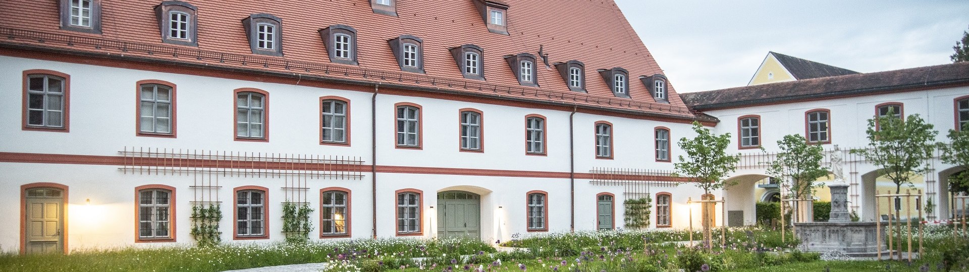 Das Bild zeigt ein großes, zweistöckiges Klostergebäude mit weißer Fassade und roten Fensterrahmen. Das steile Dach ist rot gedeckt und mit mehreren Gaubenfenstern versehen. Vor dem Gebäude liegt ein gepflegter Garten mit bunten Blumenbeeten, kleinen Bäumen und geschwungenen Kieswegen. Auf der rechten Seite führt ein Torbogen in einen weiteren Innenhof mit einem Brunnen. Die Szene ist in weiches Abendlicht getaucht, einige Fenster und Außenlampen sind beleuchtet. Der Himmel ist leicht bewölkt., © Kloster Beuerberg|Foto: Christian Schmid Das Bild zeigt ein großes, zweistöckiges Klostergebäude mit weißer Fassade und roten Fensterrahmen. Das steile Dach ist rot gedeckt und mit mehreren Gaubenfenstern versehen. Vor dem Gebäude liegt ein gepflegter Garten mit bunten Blumenbeeten, kleinen Bäumen und geschwungenen Kieswegen. Auf der rechten Seite führt ein Torbogen in einen weiteren Innenhof mit einem Brunnen. Die Szene ist in weiches Abendlicht getaucht, einige Fenster und Außenlampen sind beleuchtet. Der Himmel ist leicht bewölkt., © Kloster Beuerberg|Foto: Christian Schmid