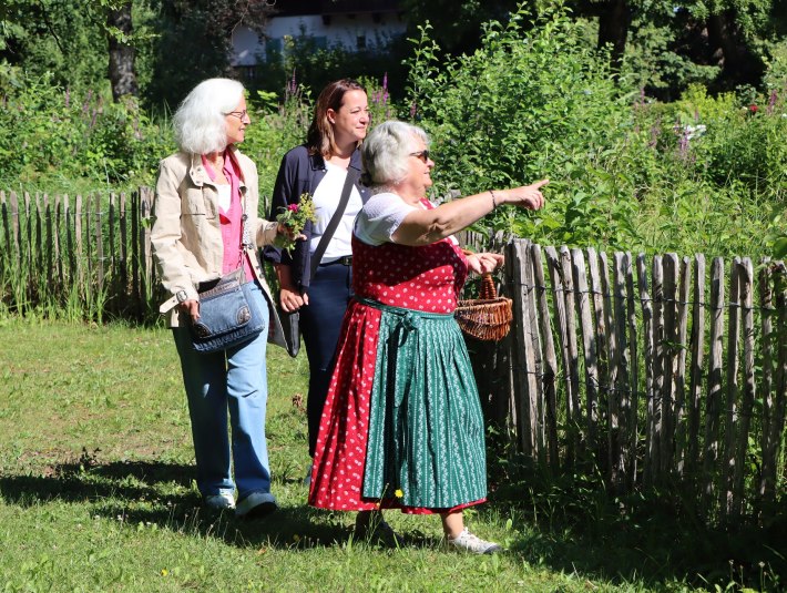 Das Bild zeigt eine Kräuterführung im Kräuter-Erlebnis-Park Bad Heilbrunn: Drei Frauen gehen an einem Gartenzaun entlang. Die Kräuterpädagogin im roten Dirndl zeigt auf etwas, während die anderen beiden Teilnehmerinnen der Kräuterführung sie ansehen., © Tölzer Land Tourismus|J. Kirschenhofer Das Bild zeigt eine Kräuterführung im Kräuter-Erlebnis-Park Bad Heilbrunn: Drei Frauen gehen an einem Gartenzaun entlang. Die Kräuterpädagogin im roten Dirndl zeigt auf etwas, während die anderen beiden Teilnehmerinnen der Kräuterführung sie ansehen., © Tölzer Land Tourismus|J. Kirschenhofer