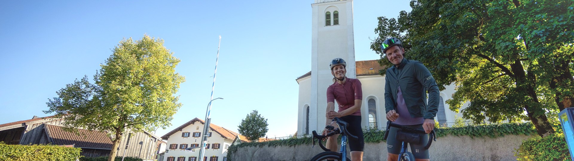 Zwei Fahrradfahrer pausieren vor der barocken St.Benediktkirche in Geretsried Gelting mit seinem markanten Zweibelturm, © Stadt Geretsried|Farmhouse 7 Zwei Fahrradfahrer pausieren vor der barocken St.Benediktkirche in Geretsried Gelting mit seinem markanten Zweibelturm, © Stadt Geretsried|Farmhouse 7