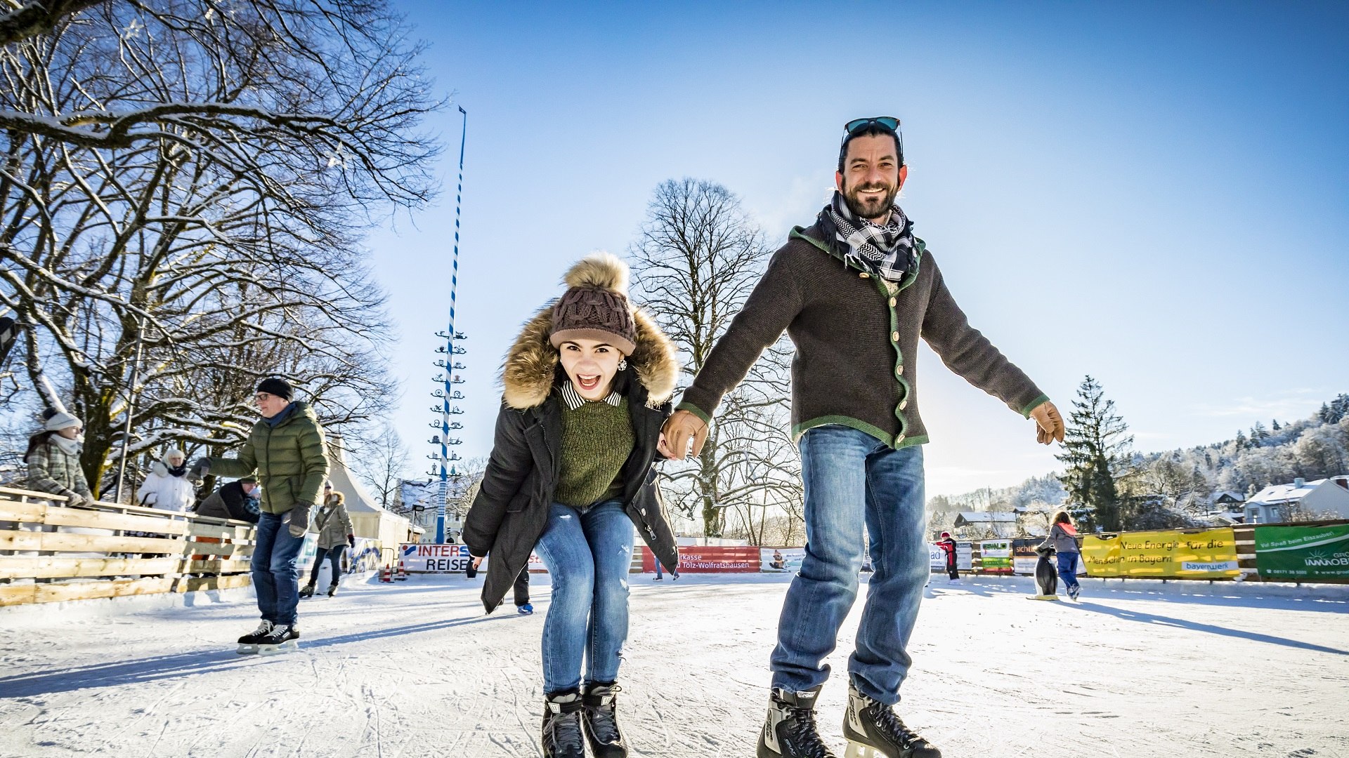 Auf der Eisfläche von Wolfratshausen laufen fröhliche Menschen mit Schlittschuhen über das Eis., © Stadt Wolfratshausen | Adrian Greiter Auf der Eisfläche von Wolfratshausen laufen fröhliche Menschen mit Schlittschuhen über das Eis., © Stadt Wolfratshausen | Adrian Greiter