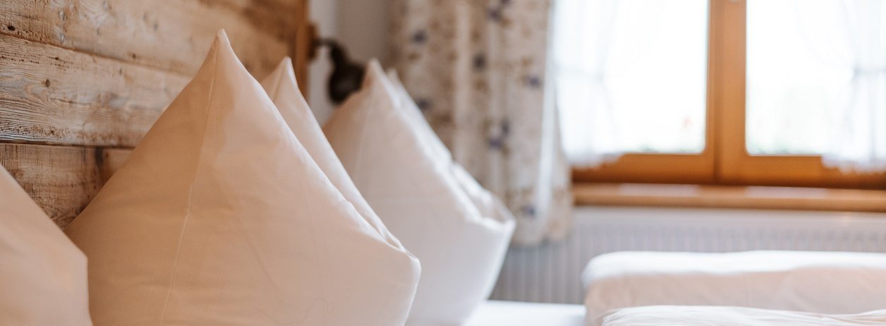 Ihre Gastgeber heißen Sie herzlich willkommen.
Das Bild zeigt zwei sorgfältig aufgeschüttelte Kopfkissen auf einem Bett mit Holzkopfteil vor dem Fenster mit geblümten Vorhängen., © Tölzer Land Tourismus | Foto: Leonie Lorenz
