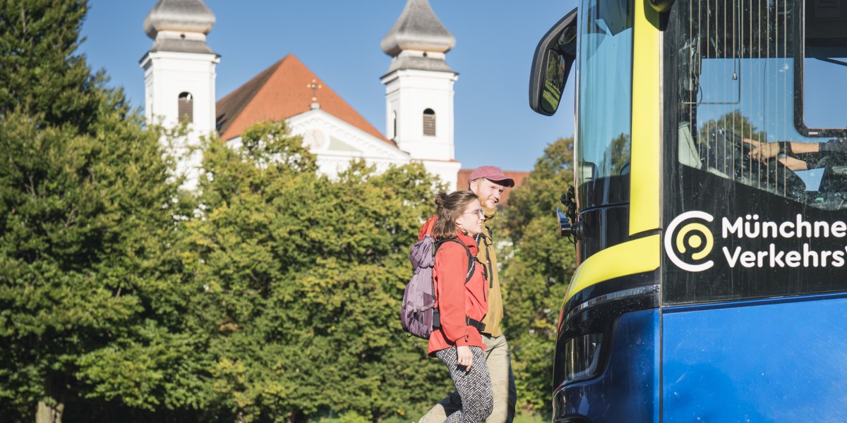 Zwei Wanderer mit Rucks&auml;cken gehen bei sonnigem Wetter auf einen blauen Bus der M&uuml;nchner Verkehrsgesellschaft zu, im Hintergrund stehen gr&uuml;ne B&auml;ume und die Klosterkirche von Schlehdorf Kirche mit zwei T&uuml;rmen., &copy; Landratsamt Bad T&ouml;lz-Wolfratshausen|Dietmar Denger