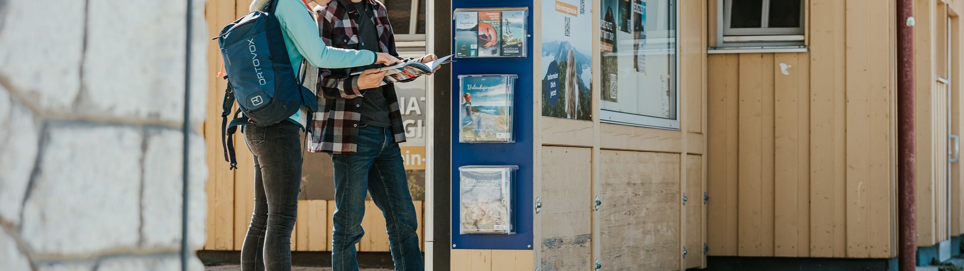 Der perfekte Urlaub beginnt schon mit der Planung der Anreise. Das Bild zeigt ein Paar, das sich am Bahnhof an einer Informationsstele informiert., © oberbayern.de|A. Köhler