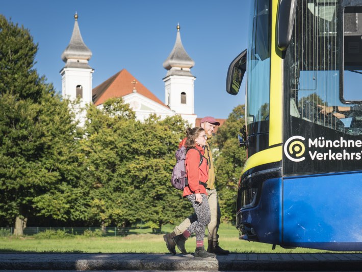 Zwei Wanderer mit Rucks&auml;cken gehen bei sonnigem Wetter auf einen blauen Bus der M&uuml;nchner Verkehrsgesellschaft zu, im Hintergrund stehen gr&uuml;ne B&auml;ume und die Klosterkirche von Schlehdorf Kirche mit zwei T&uuml;rmen., &copy; Landratsamt Bad T&ouml;lz-Wolfratshausen|Dietmar Denger
