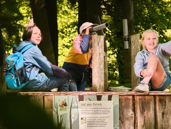 Die drei Kinder an der Station "Auf der Pirsch" im Wolfratshauser Bergwald entdecken mit dem Fernrohr Tierbilder im Wald., &copy; Stadt Wolfratshausen