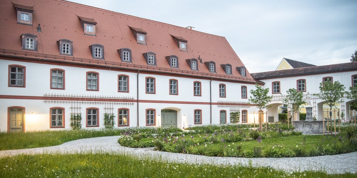 Das Bild zeigt ein gro&szlig;es, zweist&ouml;ckiges Klostergeb&auml;ude mit wei&szlig;er Fassade und roten Fensterrahmen. Das steile Dach ist rot gedeckt und mit mehreren Gaubenfenstern versehen. Vor dem Geb&auml;ude liegt ein gepflegter Garten mit bunten Blumenbeeten, kleinen B&auml;umen und geschwungenen Kieswegen. Auf der rechten Seite f&uuml;hrt ein Torbogen in einen weiteren Innenhof mit einem Brunnen. Die Szene ist in weiches Abendlicht getaucht, einige Fenster und Au&szlig;enlampen sind beleuchtet. Der Himmel ist leicht bew&ouml;lkt., &copy; Kloster Beuerberg|Foto: Christian Schmid