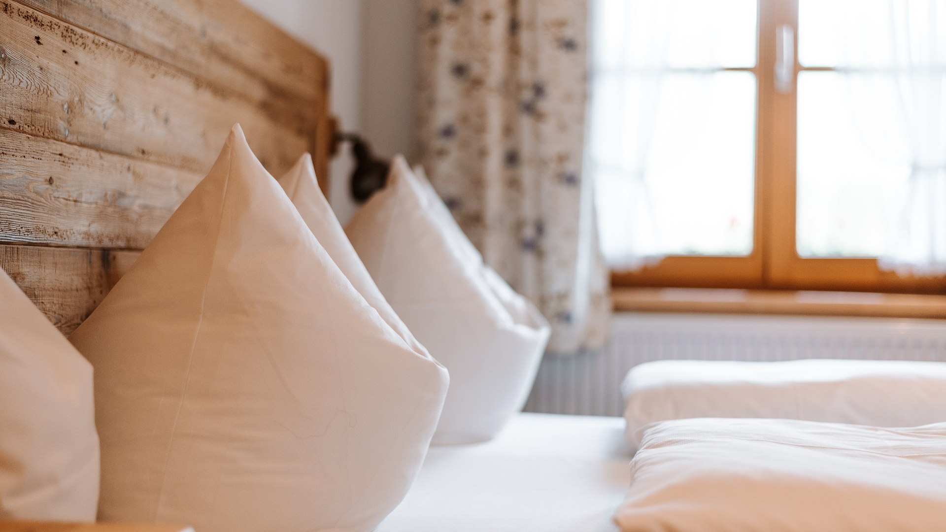Ihre Gastgeber hei&szlig;en Sie herzlich willkommen.
Das Bild zeigt zwei sorgf&auml;ltig aufgesch&uuml;ttelte Kopfkissen auf einem Bett mit Holzkopfteil vor dem Fenster mit gebl&uuml;mten Vorh&auml;ngen., &copy; T&ouml;lzer Land Tourismus | Foto: Leonie Lorenz
