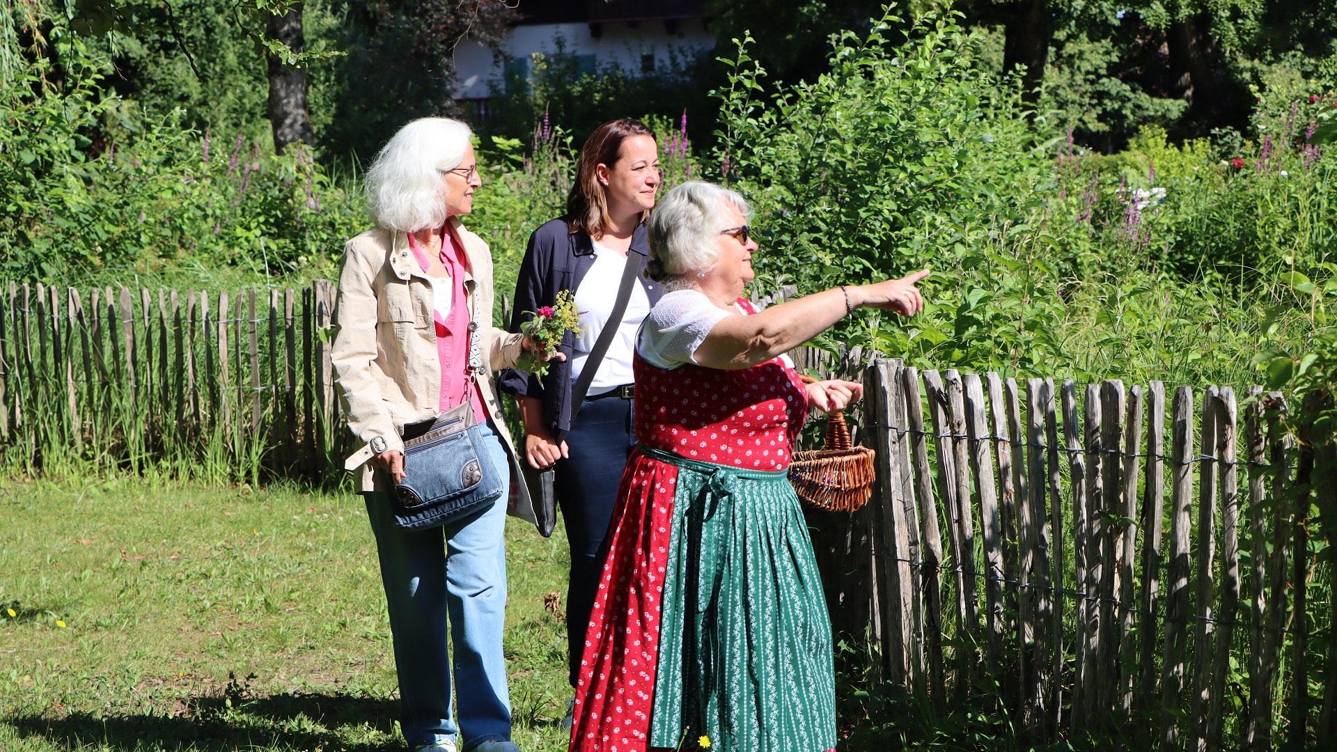 Das Bild zeigt eine Kr&auml;uterf&uuml;hrung im Kr&auml;uter-Erlebnis-Park Bad Heilbrunn: Drei Frauen gehen an einem Gartenzaun entlang. Die Kr&auml;uterp&auml;dagogin im roten Dirndl zeigt auf etwas, w&auml;hrend die anderen beiden Teilnehmerinnen der Kr&auml;uterf&uuml;hrung sie ansehen., &copy; T&ouml;lzer Land Tourismus|J. Kirschenhofer