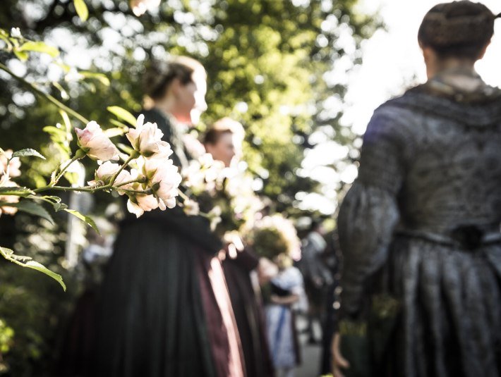 Fest verankert im Brauchtumskalender: der Kochler heimattag.
Das Bild zeigt im Vordergrund einen zwart rosa bl&uuml;henden Rosenbusch. Im Hintergrund sind unscharf Frauen im festlichen Kirchengewand der Trachtlerinnen zu sehen., &copy; Tourist Information Kochel/Walchensee|Th. Kujat
