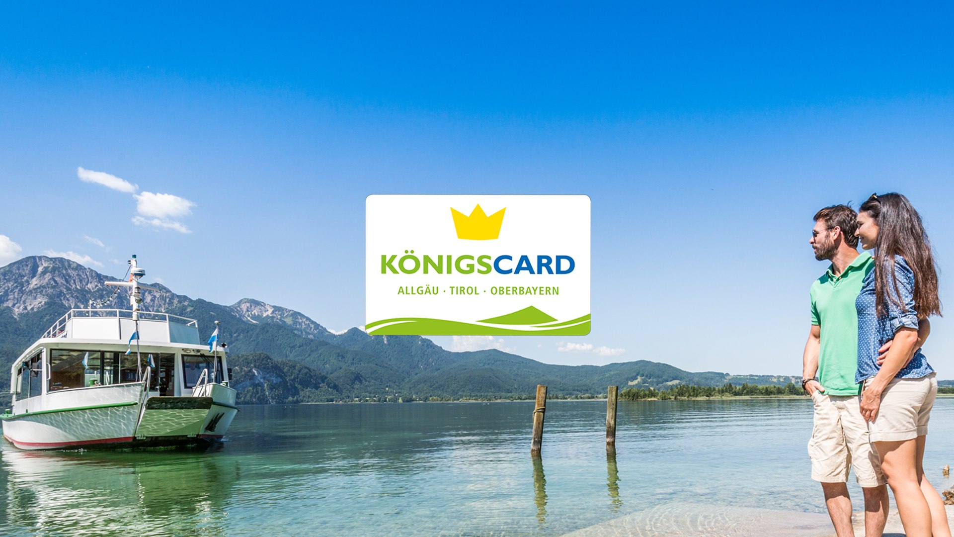 Die K&ouml;nigscard - bei ausgew&auml;hlten Gastgebern erh&auml;ltlich f&uuml;r ein MEHR an Urlaubsspa&szlig;. Das Bild zeigt ein auf das Kochelseeschiff wartende P&auml;rchen am Steg mit dem Logo der K&ouml;nigscard G&auml;stekarte, &copy; Foto: TI Kochel a. See|Thomas Kujat