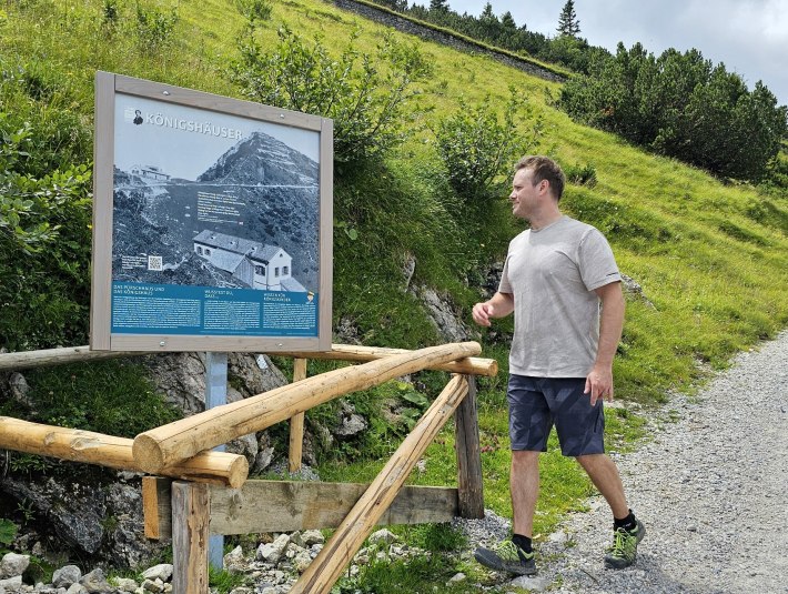 Ein Mann Mann in grauem T-Shirt und blauen Shorts liest eine Infotafel zum Themenweg &quot;König der Berge&quot; an einem Wanderweg, umgeben von grüner Natur und Hügeln des Herzogstands., © Tourist Information Kochel a. See|Foto: Daniel Weickel