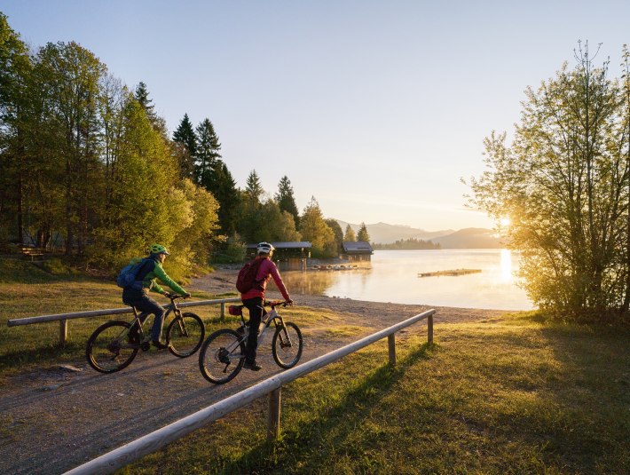 Welch Ausblick auf den Walchensee! Zwei Fahrradfahrer fahren auf den in morgendlichen Sonnenlicht getauchten Walchensee zu., &copy; Archiv T&ouml;lzer Land Tourismus| Foto: Bernd Ritschel