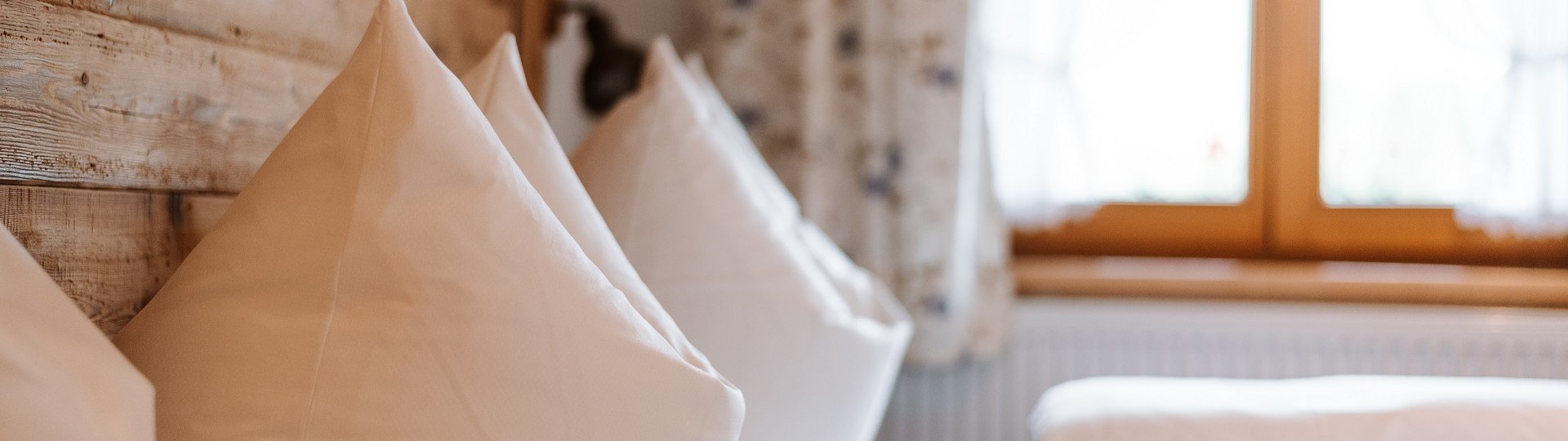 Ihre Gastgeber heißen Sie herzlich willkommen.
Das Bild zeigt zwei sorgfältig aufgeschüttelte Kopfkissen auf einem Bett mit Holzkopfteil vor dem Fenster mit geblümten Vorhängen., © Tölzer Land Tourismus | Foto: Leonie Lorenz