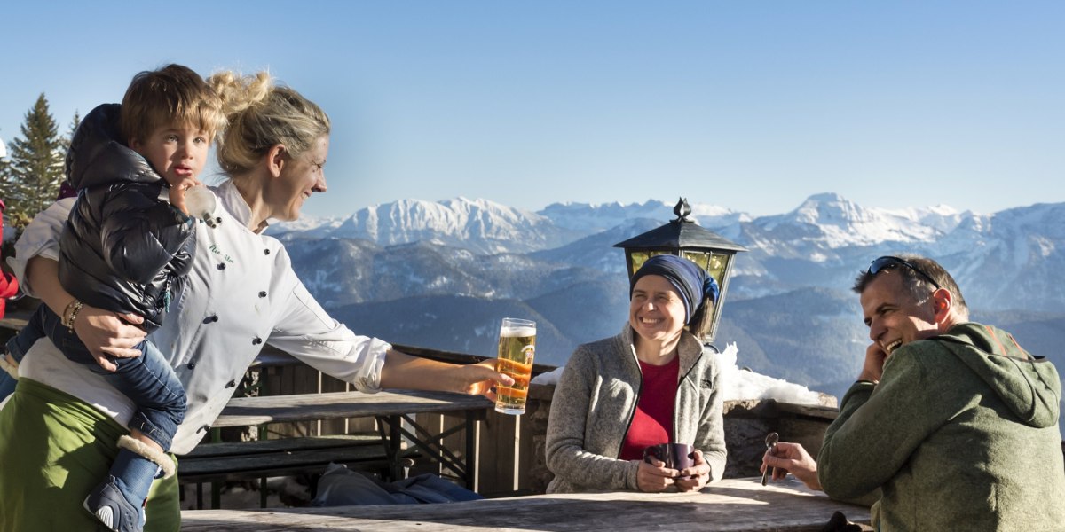 Vor der winterlichen Bergkulisse sitzen Gäste auf der Terrasse einer Berghütte. Die Gastwirtin serviert ein Getränk., © oberbayern.de|Peter von Felbert