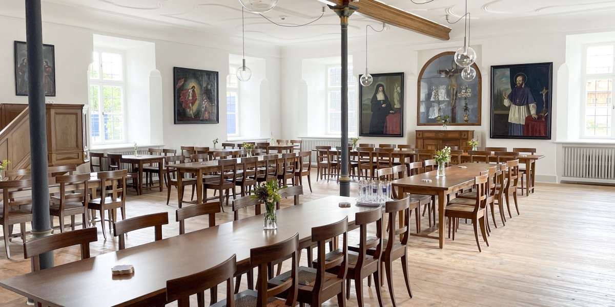 Hell und freundlich - der Restaurantbereich im Kloster Beuerberg.
Das Bild zeigt einen hellen Speisesaal mit Holzboden und langen Holztischen, umgeben von vielen St&uuml;hlen. Auf den Tischen stehen Vasen mit frischen Blumen. Gro&szlig;e Fenster lassen Tageslicht herein. An den wei&szlig;en W&auml;nden h&auml;ngen vier gro&szlig;e Gem&auml;lde mit religi&ouml;sen Motiven., &copy; Kloster Beuerberg|Foto: Christian Schmid