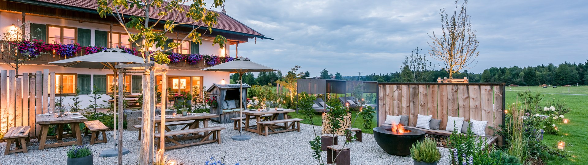 Das Bild zeigt den Außenbereich mit des Urlaub auf dem Bauernhof - Anbieters in abendlicher Stimmung mit Sonnenschirmen und Gartenlounge-Stühlen und eine große, runde Metall-Feuerstelle, © Hussnhof Foto: Günter Standl
