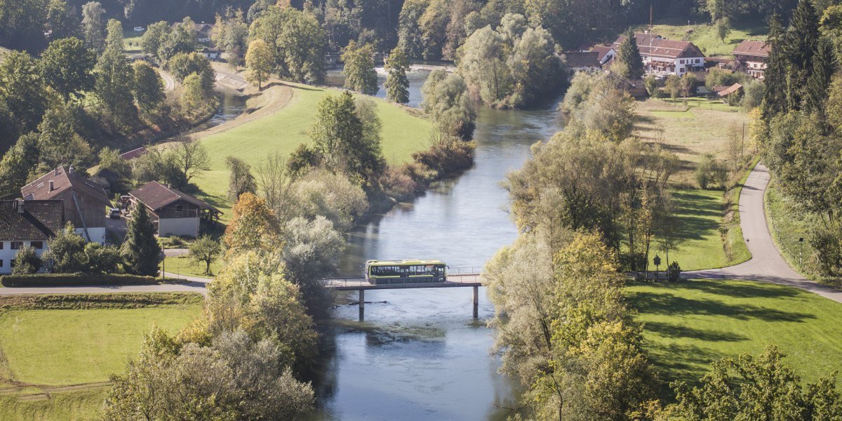 Die Loisach flie&szlig;t zwischen gr&uuml;nen Wiese und B&auml;umen, auf der Br&uuml;cke &uuml;ber den Fluss ist ein Bus zusehen; im Hintergrund zwei barocke Kircht&uuml;rme von Beuerberg und Berge unter blauem Himmel an einem sonnigen Herbsttag., &copy; Landratsamt Bad T&ouml;lz-Wolfratshausen|Dietmar Denger