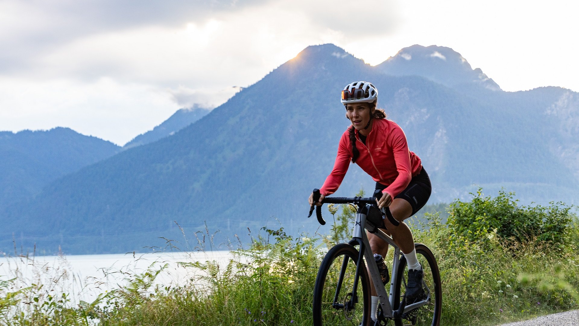 Eine Fahrradfahrerin in roter Fahrradbekleidung fährt  mit dem Gravel-Bikean einem See vor einer Bergkulisse. Der Himmel ist in sanftes Licht der Abendstimmung getaucht., © Tölzer Land Tourismus|Farmhouse7