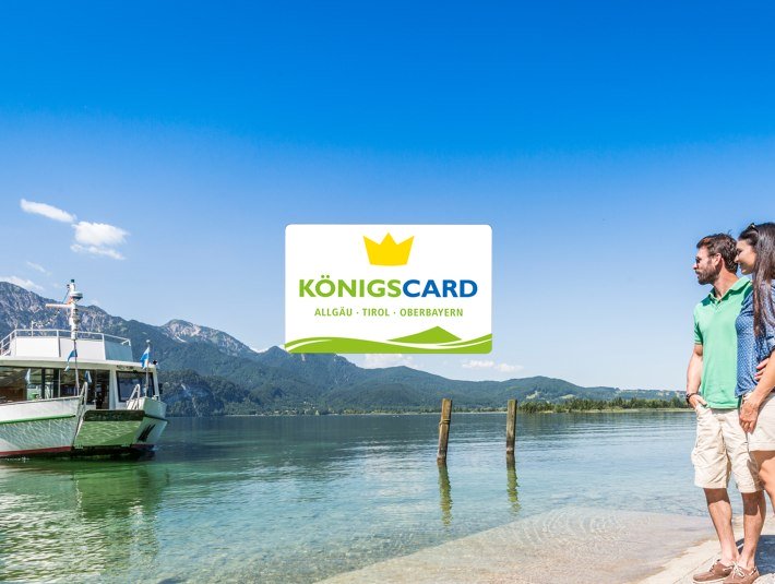 Die K&ouml;nigscard - bei ausgew&auml;hlten Gastgebern erh&auml;ltlich f&uuml;r ein MEHR an Urlaubsspa&szlig;. Das Bild zeigt ein auf das Kochelseeschiff wartende P&auml;rchen am Steg mit dem Logo der K&ouml;nigscard G&auml;stekarte, &copy; Foto: TI Kochel a. See|Thomas Kujat