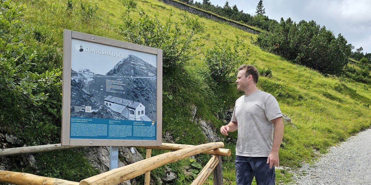 Ein Mann Mann in grauem T-Shirt und blauen Shorts liest eine Infotafel zum Themenweg "K&ouml;nig der Berge" an einem Wanderweg, umgeben von gr&uuml;ner Natur und H&uuml;geln des Herzogstands., &copy; Tourist Information Kochel a. See|Foto: Daniel Weickel