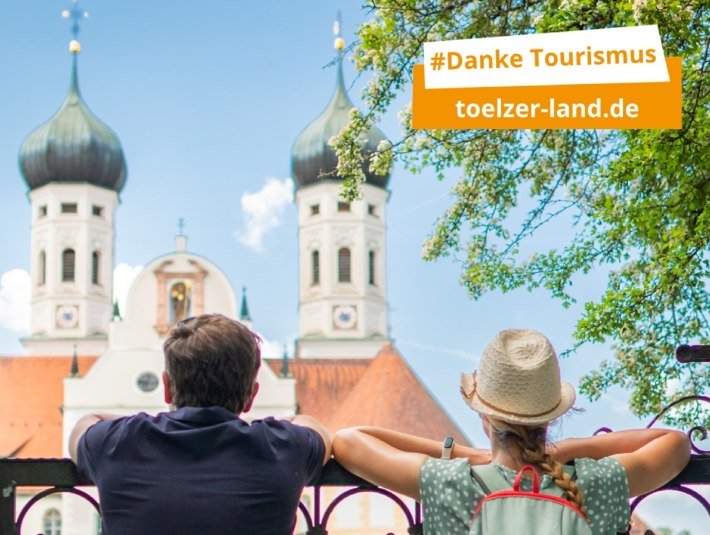 Das Bild zeigt eines der Plakate der Kampagne &quot;#Danke Tourismus&quot; und zeigt ein Paar im Sommer, das an einem schmiedeeisernen Tor lehnt und auf die Basilika des Klosters Benediktbeuern blickt., © Tölzer Land Tourismus|Foto: D. Denger