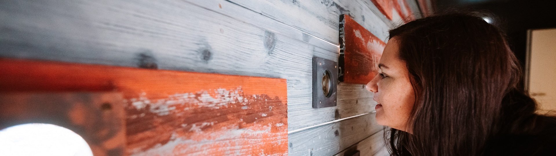 Das Bild wurde im Museum der Stadt Geretsried aufgenommen und zeigt: Eine Frau mit dunklen, langen Haaren schaut auf eine Holz-Wand mit roten Akzenten, die an einer Wand im Innenraum angebracht ist., © Tölzer Land Tourismus|Leonie Lorenz