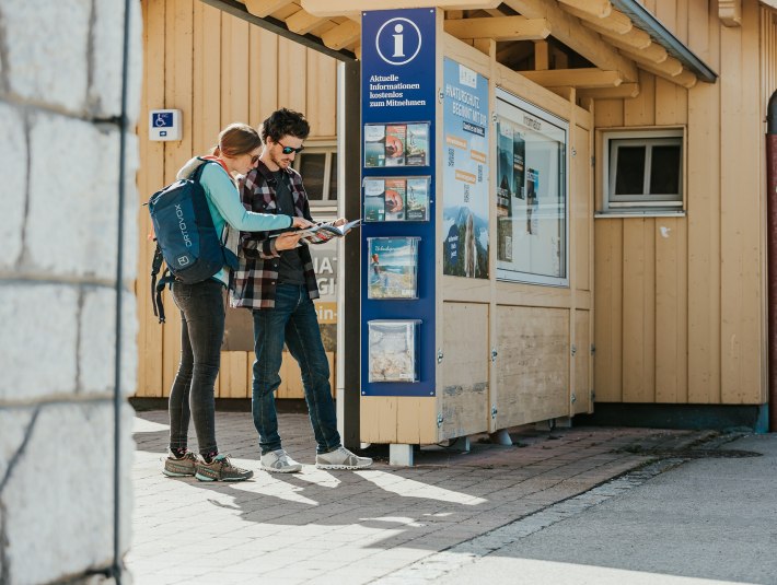 Der perfekte Urlaub beginnt schon mit der Planung der Anreise. Das Bild zeigt ein Paar, das sich am Bahnhof an einer Informationsstele informiert., © oberbayern.de|A. Köhler