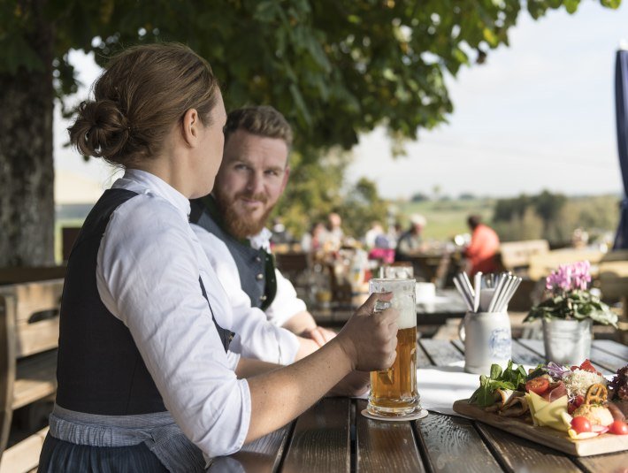 Frau und Mann sitzen in Tracht gekleidet in einem Biergarten an einem Tisch und stoßen mit ihren Getränken an, © Peter von Felbert