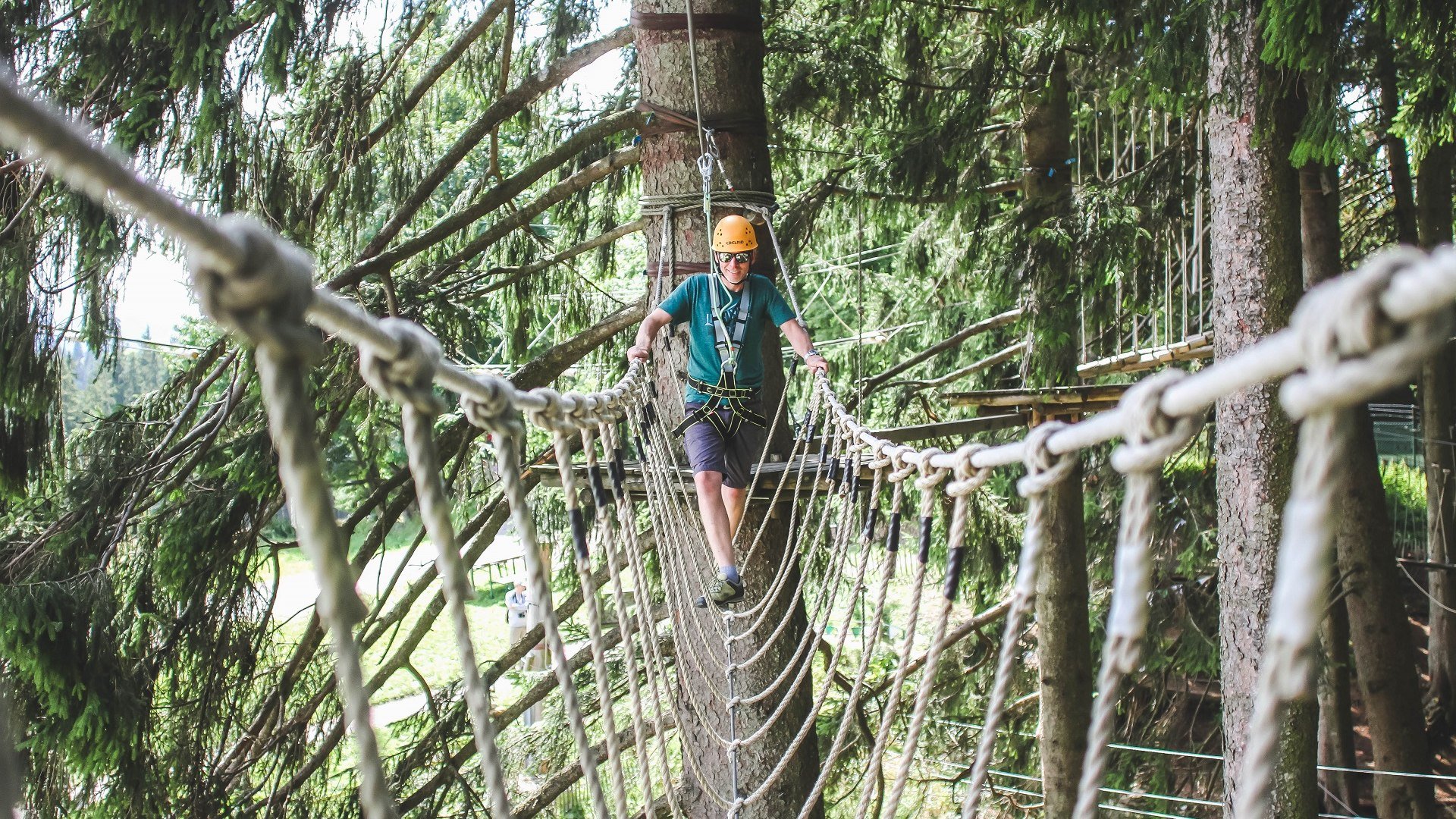 Sorgt f&uuml;r einen Adrenalin-Kick: Klettern im Hochseilgarten im Kletterwald Blomberg.
Das Bild zeigt: Person mit Helm und Klettergurt &uuml;berquert eine Seilbr&uuml;cke im Hochseilgarten; umgeben von dichten Nadelb&auml;umen, Plattformen und Sicherungsseilen., &copy; Stadt Bad T&ouml;lz|Saint Elmos