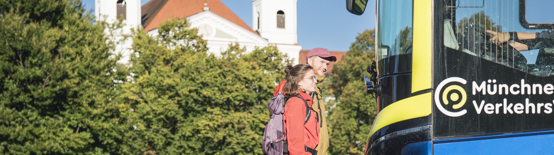 Zwei Wanderer mit Rucks&auml;cken gehen bei sonnigem Wetter auf einen blauen Bus der M&uuml;nchner Verkehrsgesellschaft zu, im Hintergrund stehen gr&uuml;ne B&auml;ume und die Klosterkirche von Schlehdorf Kirche mit zwei T&uuml;rmen., &copy; Landratsamt Bad T&ouml;lz-Wolfratshausen|Dietmar Denger