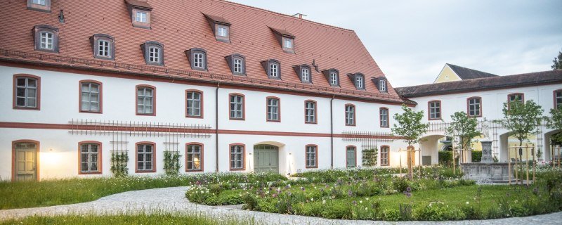 Das Bild zeigt ein großes, zweistöckiges Klostergebäude mit weißer Fassade und roten Fensterrahmen. Das steile Dach ist rot gedeckt und mit mehreren Gaubenfenstern versehen. Vor dem Gebäude liegt ein gepflegter Garten mit bunten Blumenbeeten, kleinen Bäumen und geschwungenen Kieswegen. Auf der rechten Seite führt ein Torbogen in einen weiteren Innenhof mit einem Brunnen. Die Szene ist in weiches Abendlicht getaucht, einige Fenster und Außenlampen sind beleuchtet. Der Himmel ist leicht bewölkt., © Kloster Beuerberg|Foto: Christian Schmid