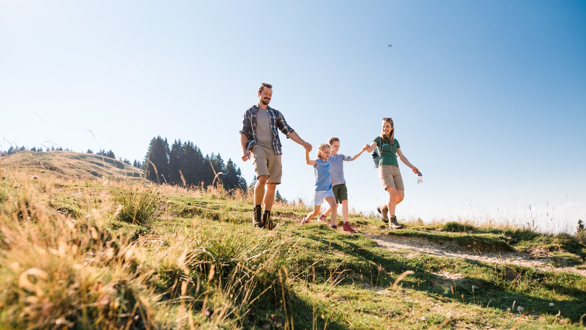Eine vierk&ouml;pfige Familie wandert Hand in Hand auf dem Wanderweg einer Almwiese, &copy; T&ouml;lzer Land Tourismus Foto: Leonie Lorenz