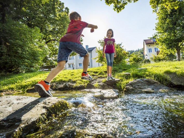 Die Fl&ouml;&szlig;erei spielerisch erleben beim Kinderfl&szlig;erpfad in Wolfratshausen. Zu sehen sind: Zwei Kinder an einem kleinen Bachlauf im Sommer. Ein Kind springt gerade &uuml;ber die Steine im Wasser., &copy; Stadt Wolfratshausen|Foto: A. Greiter