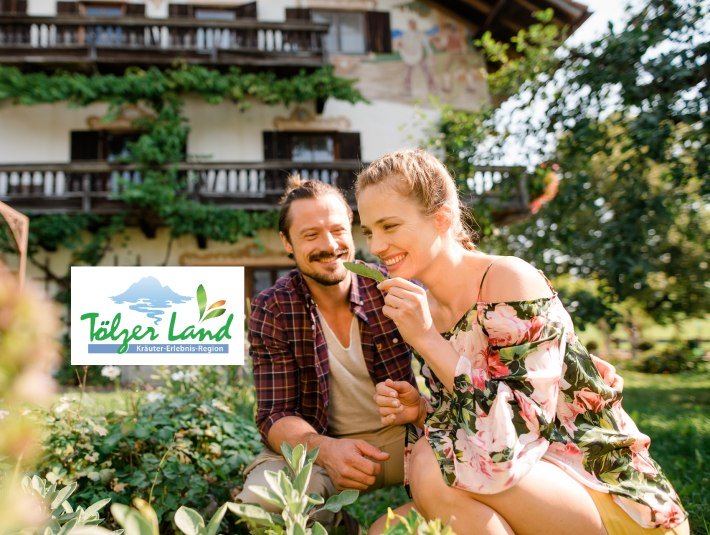 Pärchen im Garten beim Schnuppern an Kräutern mit Kräuter-Erlebnis-Logo, © Archiv Tölzer Land Tourismus|Fotograf: Leonie Lorenz