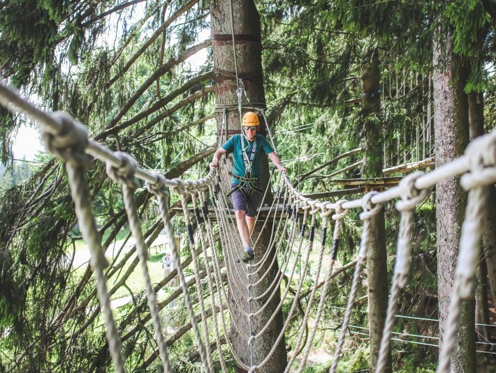Sorgt f&uuml;r einen Adrenalin-Kick: Klettern im Hochseilgarten im Kletterwald Blomberg.
Das Bild zeigt: Person mit Helm und Klettergurt &uuml;berquert eine Seilbr&uuml;cke im Hochseilgarten; umgeben von dichten Nadelb&auml;umen, Plattformen und Sicherungsseilen., &copy; Stadt Bad T&ouml;lz|Saint Elmos