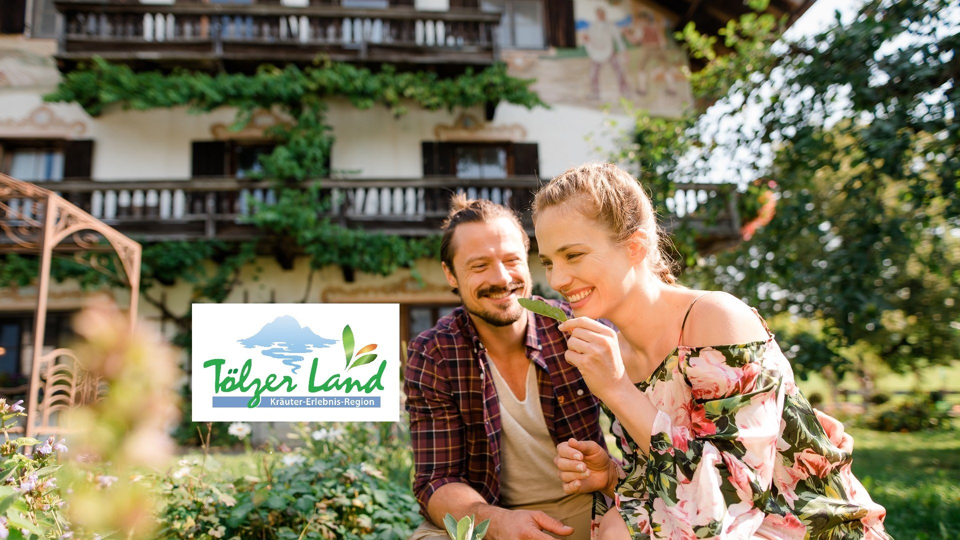 Pärchen im Garten beim Schnuppern an Kräutern mit Kräuter-Erlebnis-Logo, © Archiv Tölzer Land Tourismus|Fotograf: Leonie Lorenz