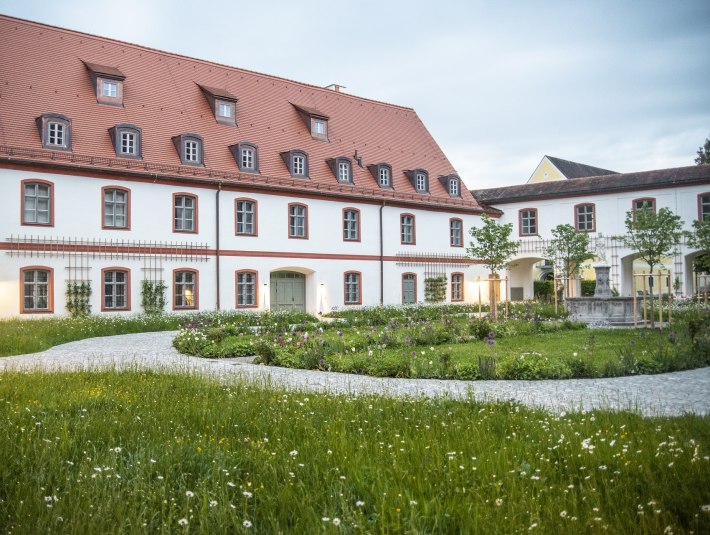 Das Bild zeigt ein großes, zweistöckiges Klostergebäude mit weißer Fassade und roten Fensterrahmen. Das steile Dach ist rot gedeckt und mit mehreren Gaubenfenstern versehen. Vor dem Gebäude liegt ein gepflegter Garten mit bunten Blumenbeeten, kleinen Bäumen und geschwungenen Kieswegen. Auf der rechten Seite führt ein Torbogen in einen weiteren Innenhof mit einem Brunnen. Die Szene ist in weiches Abendlicht getaucht, einige Fenster und Außenlampen sind beleuchtet. Der Himmel ist leicht bewölkt., © Kloster Beuerberg|Foto: Christian Schmid
