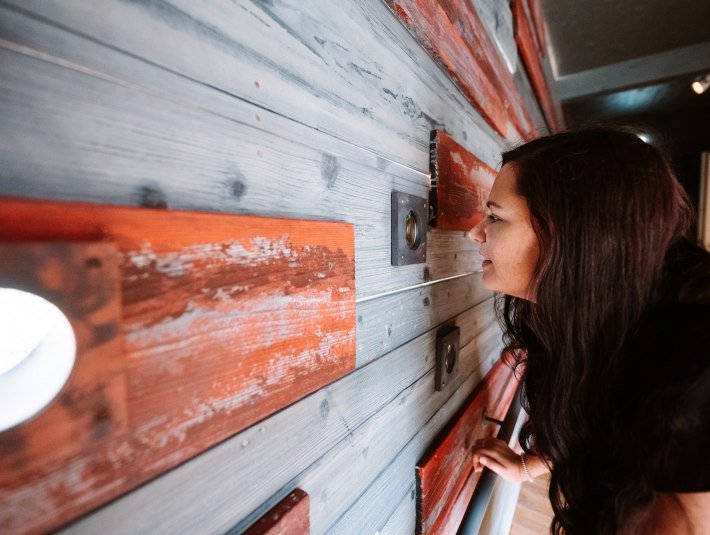 Das Bild wurde im Museum der Stadt Geretsried aufgenommen und zeigt: Eine Frau mit dunklen, langen Haaren schaut auf eine Holz-Wand mit roten Akzenten, die an einer Wand im Innenraum angebracht ist., © Tölzer Land Tourismus|Leonie Lorenz