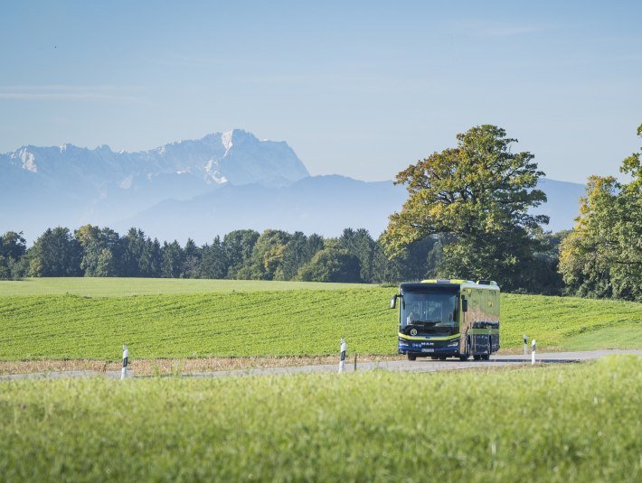ZU sehen auf dem Bild: Gr&uuml;ner Linienbus auf Landstra&szlig;e, umgeben von gr&uuml;nen Feldern und B&auml;umen; im Hintergrund die Alpenkette mit der markanten Zugspitze., &copy; Landratsamt Bad T&ouml;lz-Wolfratshausen, Foto: D. Denger