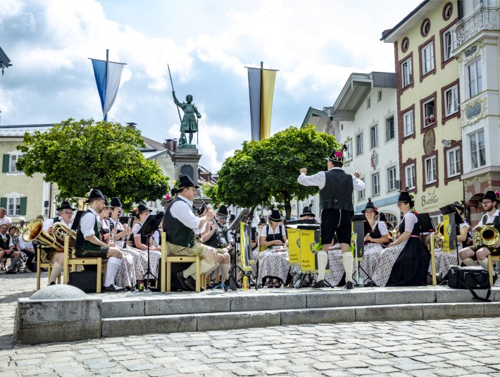 Auf dem Bild spielt die Blaskapelle auf dem Platz vor dem sog. Winzerer&quot;-Denkmal. Der Dirigent steht vorne, während Musiker auf Stühlen sitzen und musizieren. Im Hintergrund sind die mit Lüftlmalereien verzierten Gebäude der historischen Marktstraße von Bad Tölz und Bäume zu sehen., © Stadt Bad Tölz