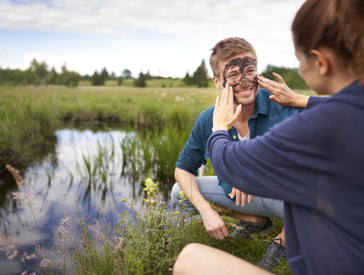 Ein vielf&auml;ltiges Veranstaltungsprogramm l&auml;sst 2025 G&auml;ste und Einheimische das Heilmittel Moor erleben. Das Bild zeigt ein junges Paar, das vor der Moorlandschaft bei Bad T&ouml;lz kniet. Dem jungen Mann wird eine Moorpackung ins Gesicht aufgetragen. , &copy; Stadt Bad T&ouml;lz
