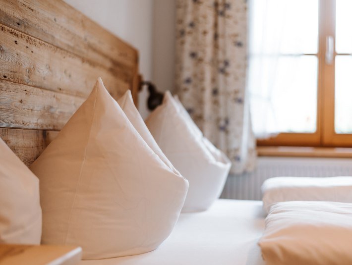 Ihre Gastgeber hei&szlig;en Sie herzlich willkommen.
Das Bild zeigt zwei sorgf&auml;ltig aufgesch&uuml;ttelte Kopfkissen auf einem Bett mit Holzkopfteil vor dem Fenster mit gebl&uuml;mten Vorh&auml;ngen., &copy; T&ouml;lzer Land Tourismus | Foto: Leonie Lorenz